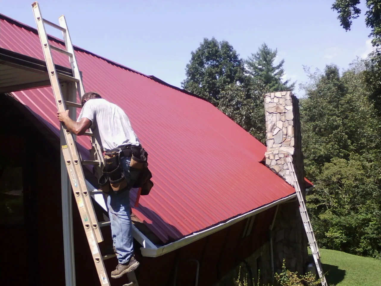 Roofer on a ladder beside a red metal roof and stone chimney in Bear